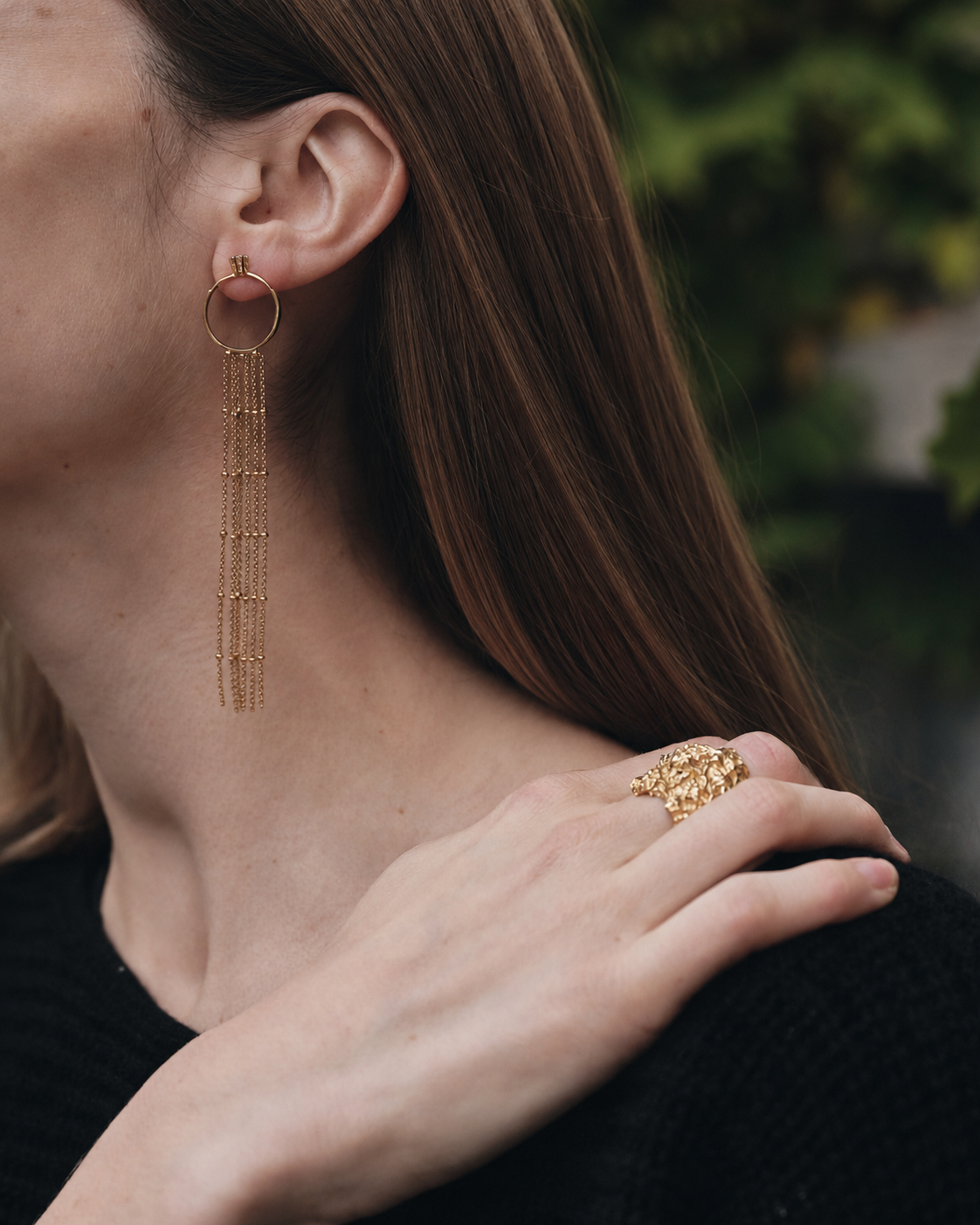 Close-up of a person wearing gold hoop earrings and ring with a blurred natural background
