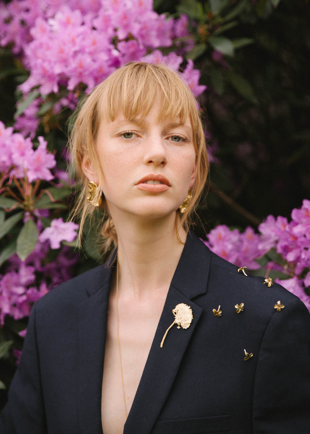 Woman wearing a black blazer with handcrafted gold lilac earrings and brooches in front of pink flowers.