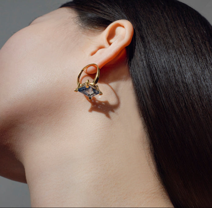 Close-up of a handcrafted gold earring with a blue gemstone on a neutral background
