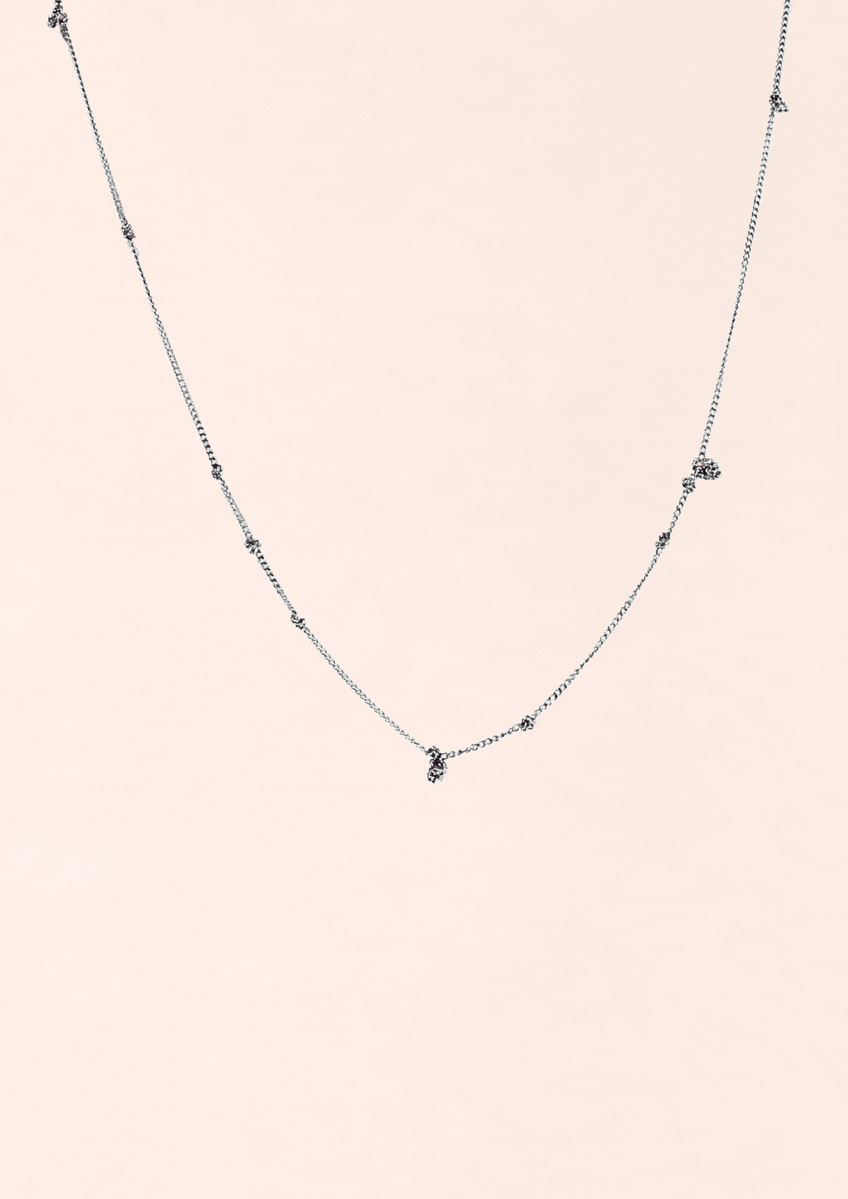 A delicate silver-toned chain necklace with small spaced bead details and a tiny central charm, displayed against a solid pale pink background.