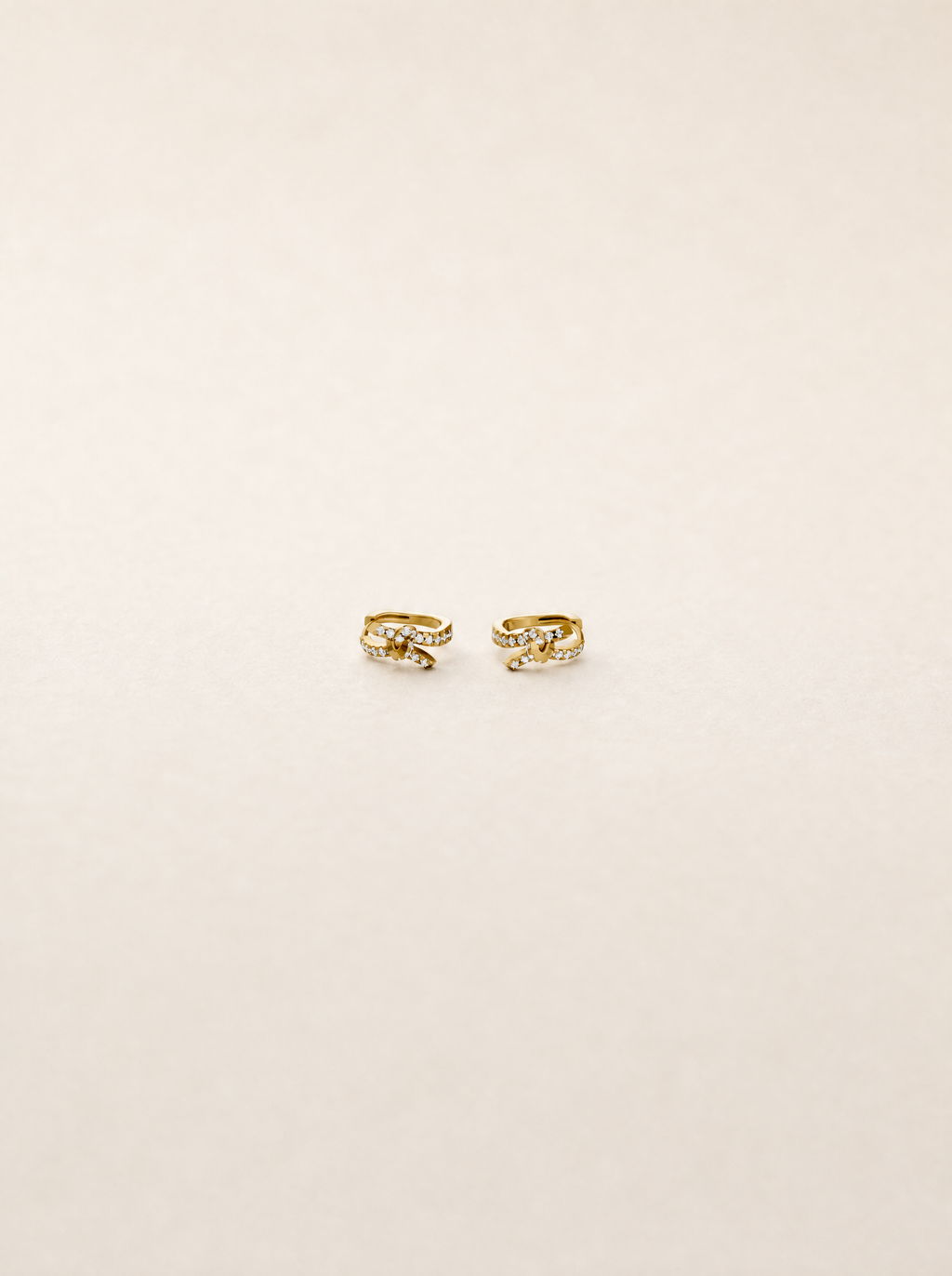 A pair of small gold huggie hoop earrings with clear gemstones, displayed on a clean, light beige background.