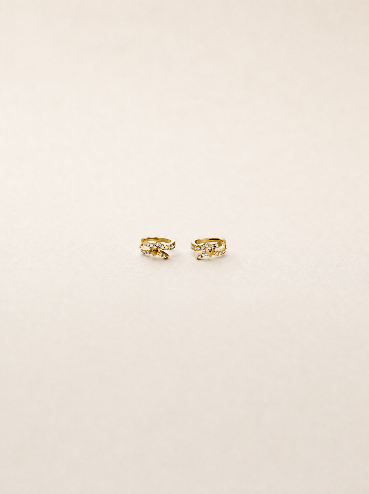 A pair of small gold huggie hoop earrings with clear gemstones, displayed on a clean, light beige background.
