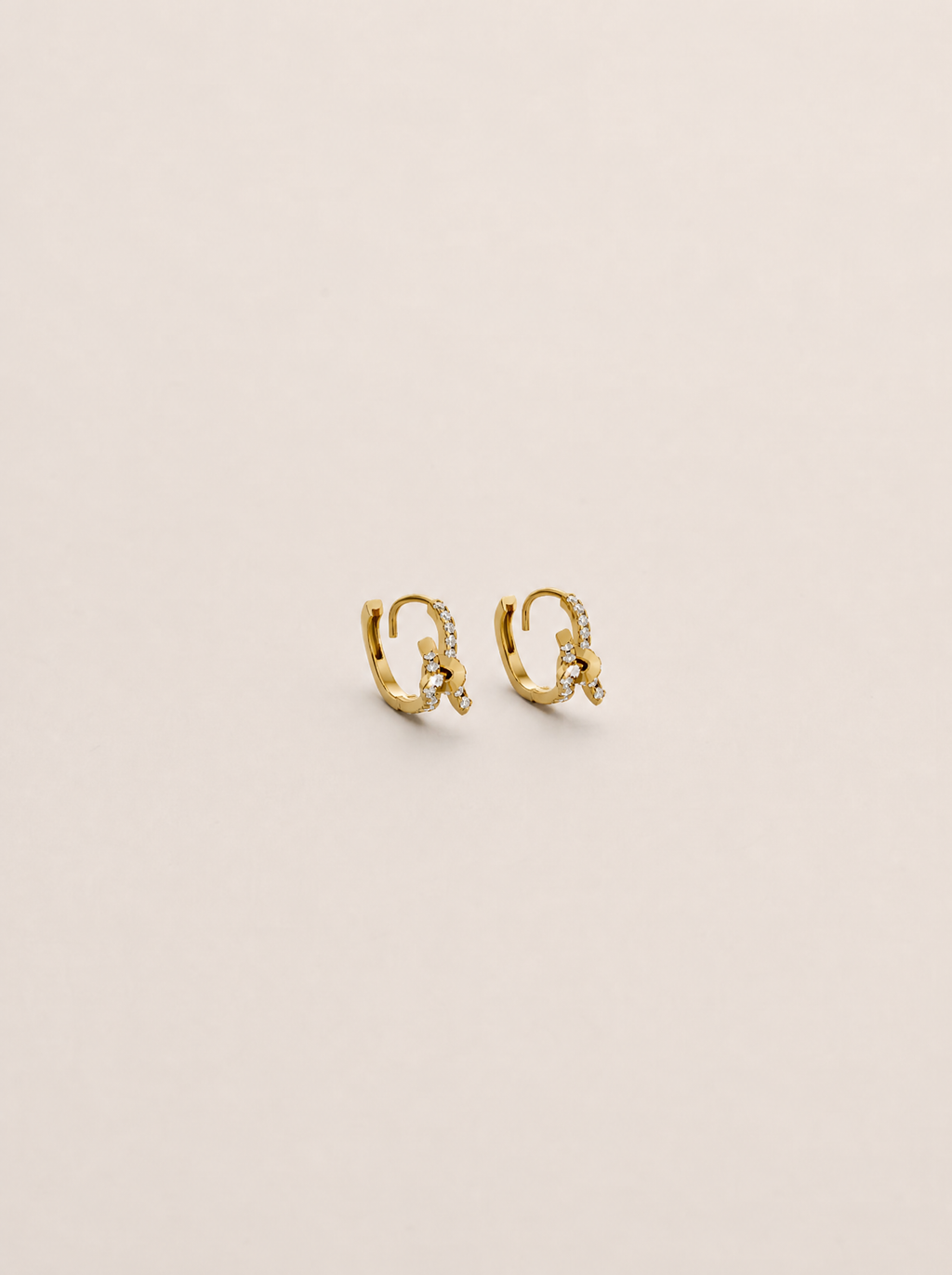 A pair of small gold huggie hoop earrings with delicate pavé-set clear stones, displayed on a clean, solid, warm off-white background.