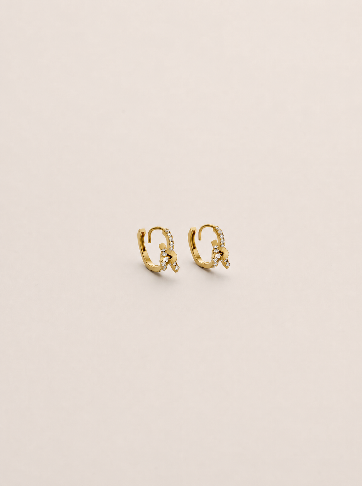 A pair of small gold huggie hoop earrings with delicate pavé-set clear stones, displayed on a clean, solid, warm off-white background.