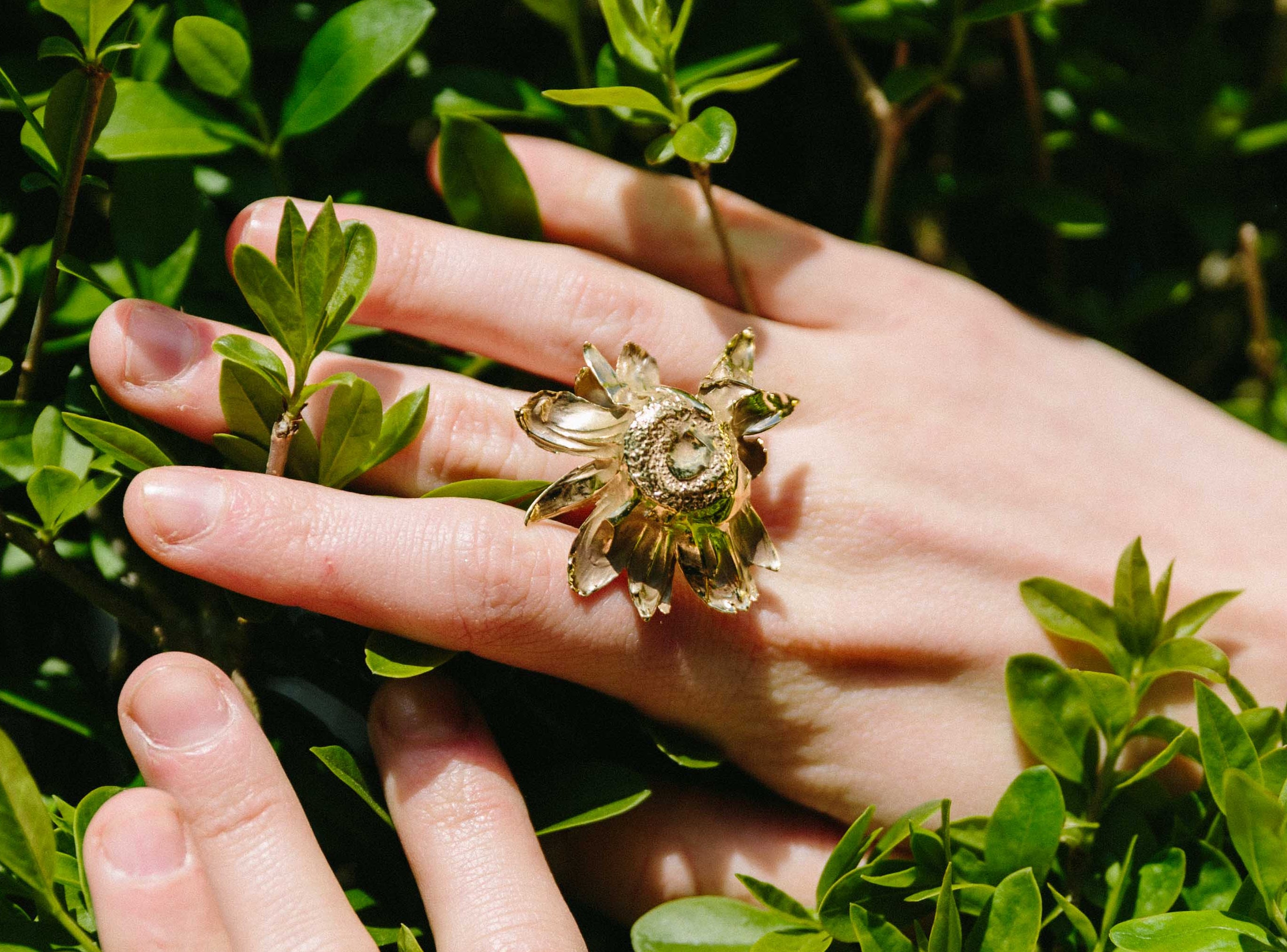 Two hands with handcrafted gold Floret ring on a green leafy background